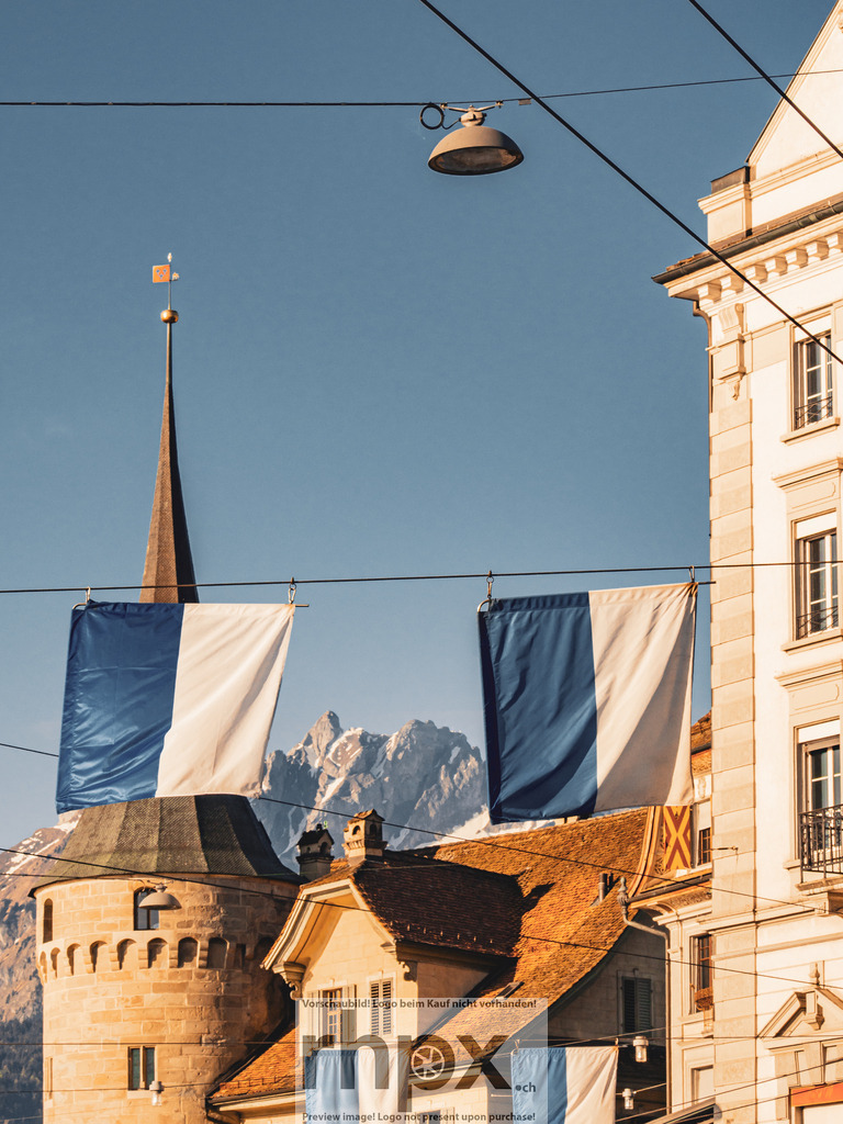 Luzern mit Pilatus und Fahnen im Morgenlicht | Luzern im klaren Morgenlicht, eingerahmt von zwei blau-weissen Fahnen. Dahinter zeichnet sich der Pilatus scharf vor dem Himmel ab.  - Realisiert mit Pictrs.com