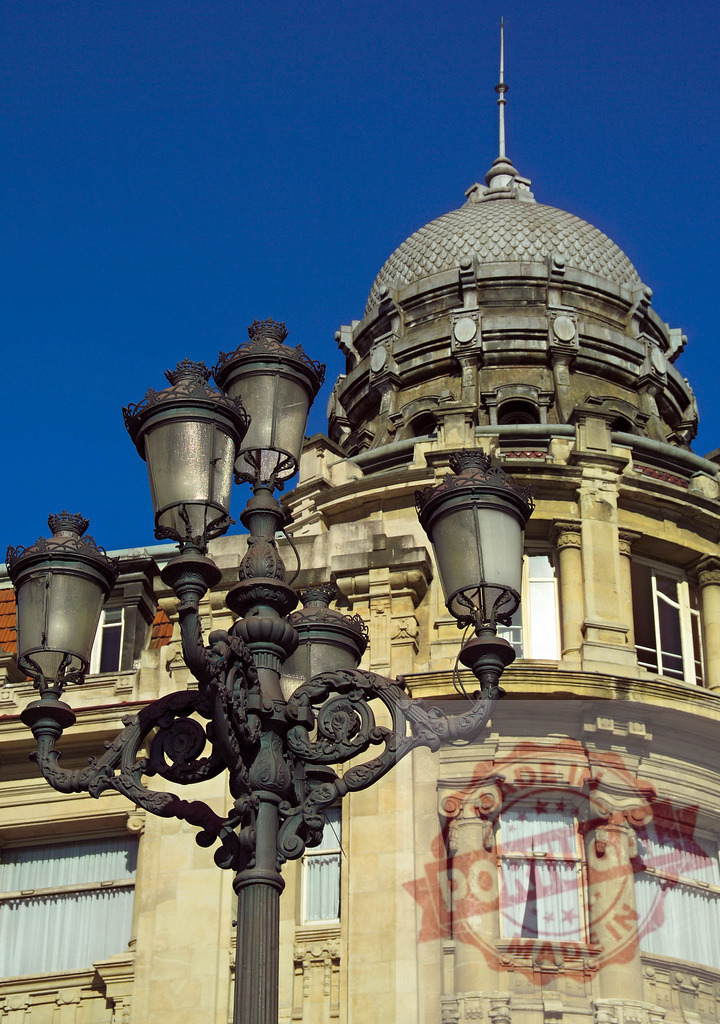 Ornate street lamp against historic architecture and clear blue sky. Spain - Bilbao | Ornate street lamp standing in front of a classical building with a dome, under a clear blue sky - Realized with Pictrs.com