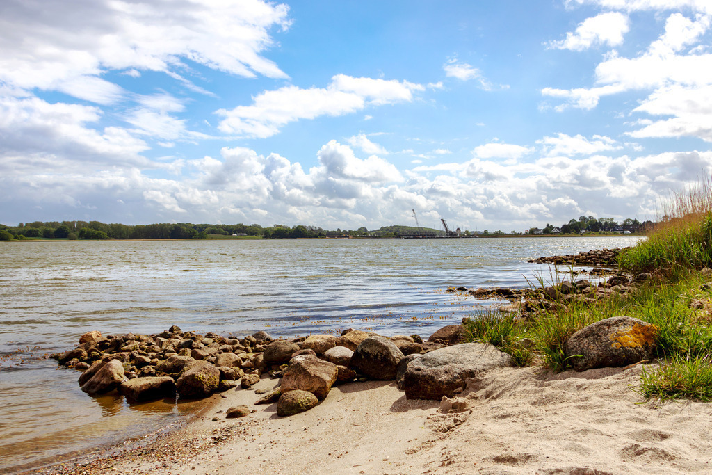 Wandbild: Schleiufer in Lindaunis im Frühling | Dieses Wandbild im Querformat zeigt den Uferbereich der Schlei im Frühling. Im Hintergrund ist die Lindaunisbrücke zu sehen. Am blauen Himmel befinden sich einige helle Wolken.  - Realisiert mit Pictrs.com