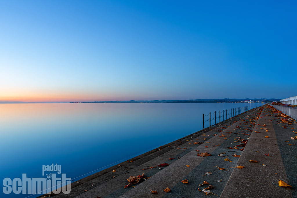 Seeufer bei Abenddämmerung bei Bregenz am Bodensee | In der sanften Abenddämmerung liegt das Bodenseeufer bei Bregenz in einer besonders ruhigen und stimmungsvollen Atmosphäre. Das letzte Licht des Tages spiegelt sich auf dem Wasser und verleiht der Landschaft eine zauberhafte, fast meditative Stimmung. - Realisiert mit Pictrs.com