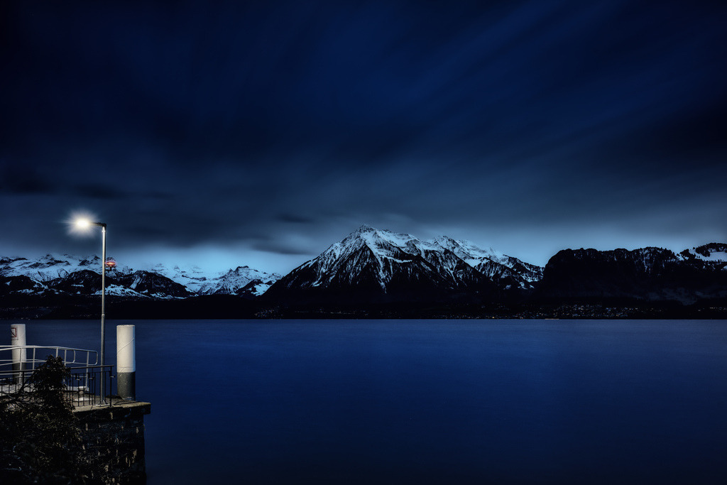 Licht in der Dunkelheit | Ein Nachtpanorama zeigt einen ruhigen See unter einem tiefblauen Himmel. Links im Vordergrund ragt eine leuchtende Strassenlaterne auf einem steinernen Pier empor, deren helles Licht einen Sterneneffekt erzeugt. Im Mittelgrund erstreckt sich die glatte, dunkelblaue Wasserfläche des Sees. Dahinter erheben sich majestätische, schneebedeckte Berge mit scharfen, dunklen Konturen gegen den Horizont. Die Bergspitzen sind in Weiss und Hellblau getaucht, während die unteren Hänge dunkel bleiben. Am Fusse der Berge sind vereinzelt kleine, gelbliche Lichter einer Siedlung erkennbar. Der Himmel ist in verschiedenen Blautönen gehalten, von einem helleren Blau über den Bergen bis zu einem fast schwarzen Dunkelblau oben, mit leichten Wolkenschleiern durch die Langzeitbelichtung. - Realisiert mit Pictrs.com
