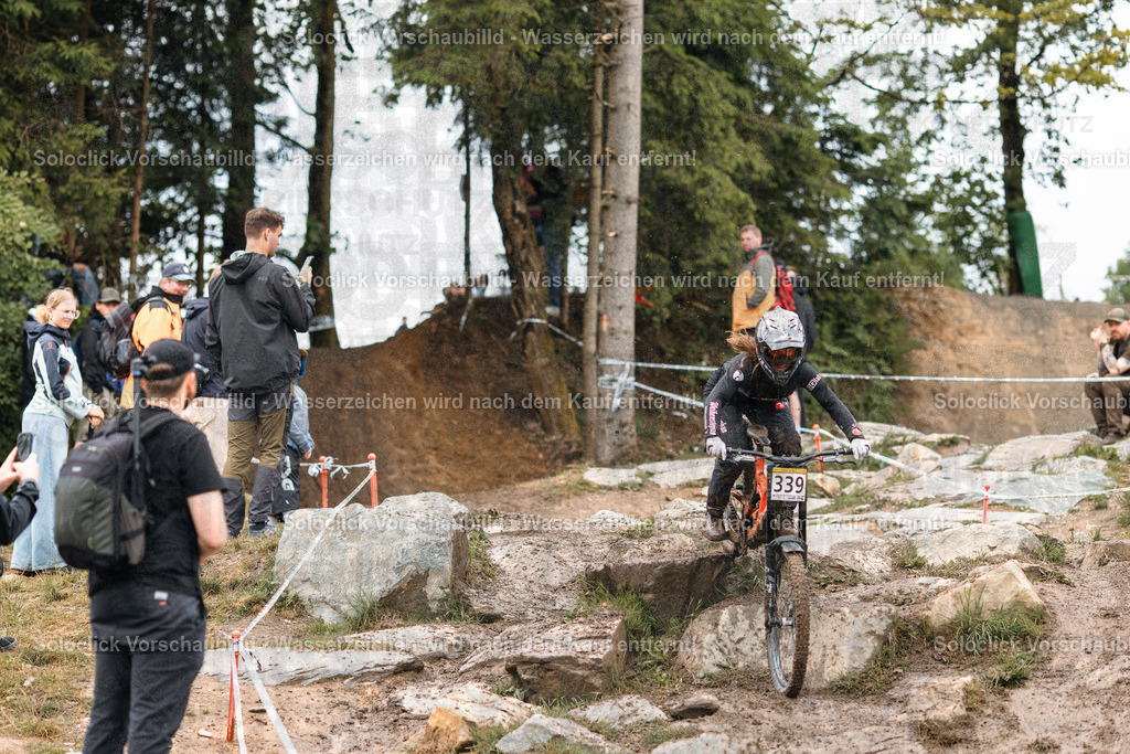 Mountainbike-Action im Wald | Das Foto zeigt einen Mountainbiker in voller Ausrüstung, der einen Sprung über eine Felswand während eines Wettkampfs oder Trainings absolviert. Der Biker trägt einen schwarzen Helm mit gelben Akzenten und eine dunkle Brille. Er trägt eine rote Jacke und schwarze Handschuhe. Auf seinem Rücken befindet sich eine Nummer 339. Der Hintergrund besteht aus dichtem Wald und einer Gruppe von Zuschauern, die den Wettkampf beobachten. Der Pfad ist staubig und mit Felsen übersät. Das Wetter scheint bewölkt zu sein. - Realisiert mit Pictrs.com