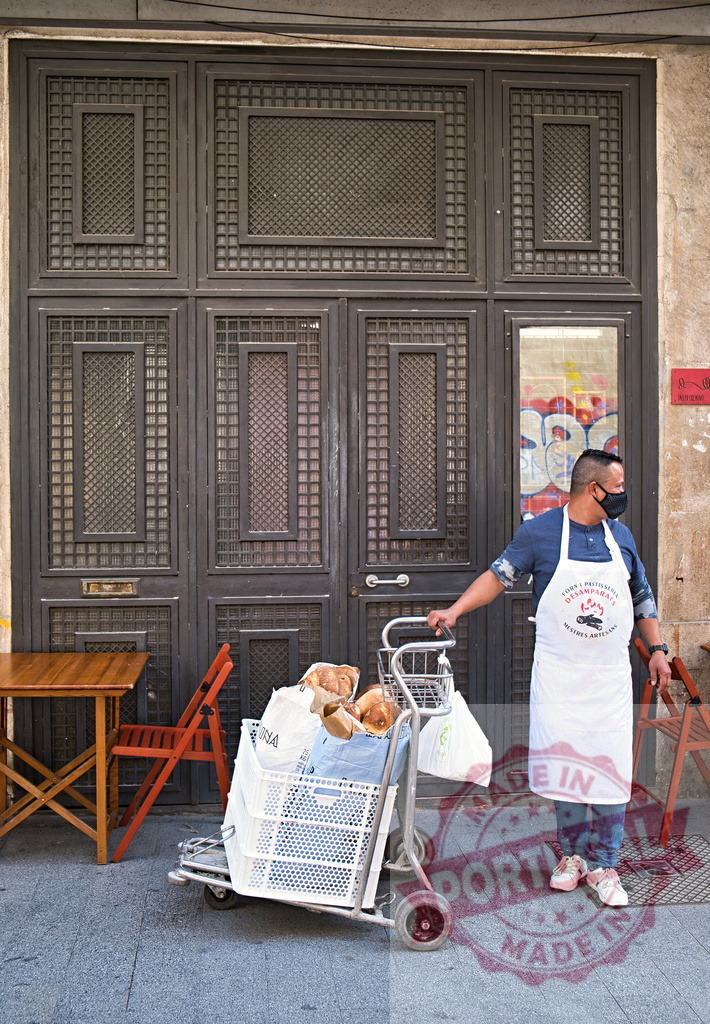 Baker wearing face mask delivering bread on Barcelona street | Baker wearing apron and face mask pushing cart filled with fresh bread for delivery - Realized with Pictrs.com