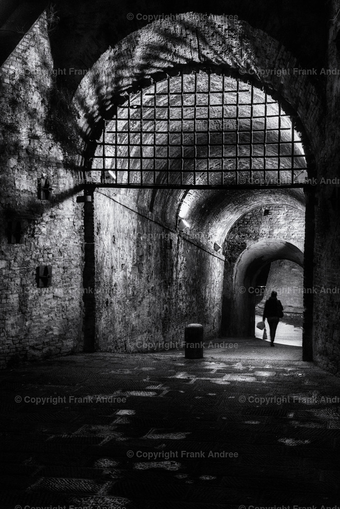 Nacht in Lucca | Eine Frau geht allein durch einen beleuchteten Gang in der Stadtmauer von Lucca Toskana. Straßenfotografie aus Italien in schwarz und weiß. - Realisiert mit Pictrs.com