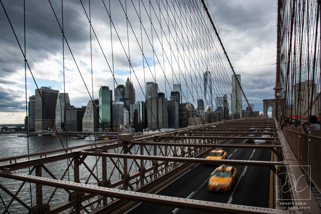 New York – Urban Motion | Brooklyn Bridge Fine Art Print | Dieses Fine-Art-Foto zeigt die Skyline von Manhattan vom Standpunkt der Brooklyn Bridge aus.Die klare Architektur des World Trade Center, die dramatischen Wolken und die bewegten gelben Taxis verbinden Ruhe und Dynamik zu einer modernen urbanen Komposition.Die Linien der Brückenkonstruktion führen den Blick tief in die Stadt und erzeugen ein Gefühl von Richtung, Raum und Energie.Ein zeitloses Statement-Piece für moderne Wohnräume, Büros, Kanzleien und Design-Interiors. - Realisiert mit Pictrs.com
