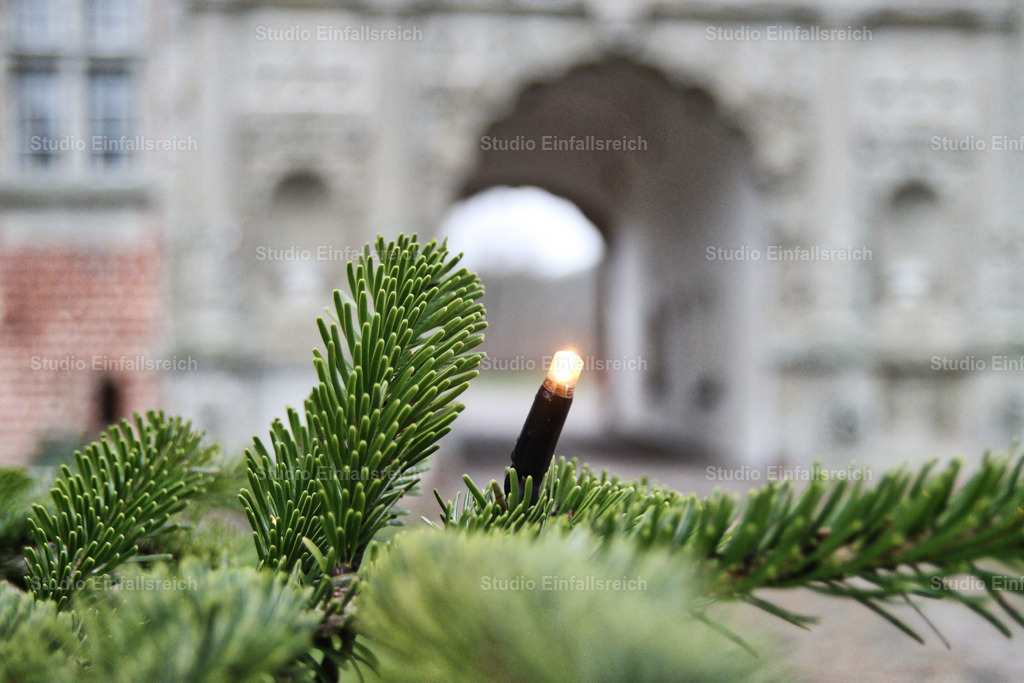 Festliches Licht am Tannenbaum vor historischer Schlosskulisse | Eine stimmungsvolle Nahaufnahme eines grünen Tannenzweigs mit einer leuchtenden LED-Lichterkette. Im sanften Bokeh des Hintergrunds ist die beeindruckende Architektur eines historischen Tores oder Schlosses zu erkennen. Das Bild vermittelt eine ruhige, weihnachtliche Atmosphäre und verbindet Natur mit klassischer Baukunst. Ideal für Weihnachtsgrüße, Reiseberichte oder saisonale Dekoration. - Realisiert mit Pictrs.com