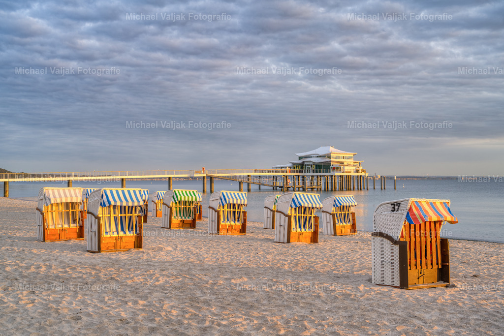 Morgensonne am Timmendorfer Strand | An einem bewölkten Morgen schafft es die Sonne kurz durch die Wolkendecke und taucht die Seeschlösschenbrücke mit dem Teehaus am Ende und die Strandkörbe am Strand in ein warmes Licht.  - Realisiert mit Pictrs.com