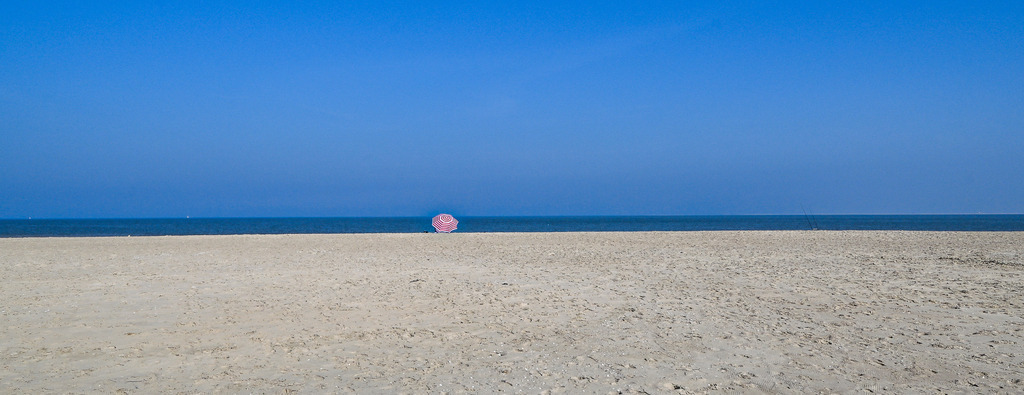 Ein Quäntchen Rot am Horizont | Ein schier endloser Sandstrand auf Texel trifft auf das tiefe Blau von Nordsee und Himmel. Mitten in dieser weiten, fast beruhigenden Einsamkeit setzt ein einzelner, rot-weiß gestreifter Sonnenschirm einen lebendigen Farbakzent. Das Bild fängt die pure Magie der Entschleunigung ein: Das sanfte Rauschen der Wellen, die frische Meeresbrise und das Gefühl, für einen Moment die Welt ganz für sich alleine zu haben. Ein Sehnsuchtsort für alle, die Stille und Weite suchen. - Realisiert mit Pictrs.com