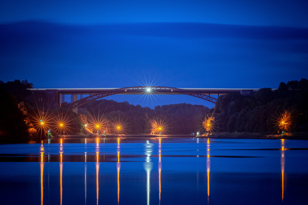 Levensauer Hochbrücke | Die Aufnahme zeigt die imposante Levensauer Hochbrücke, die majestätisch über den Nord-Ostsee-Kanal im nächtlichen Gewand thront. Die Brücke erhebt sich stolz über das dunkle, ruhige Wasser und bildet eine eindrucksvolle Verbindung zwischen den Ufern. Die Aufnahme vermittelt eine ruhige, fast meditative Atmosphäre. Die Levensauer Hochbrücke, in ihrer architektonischen Pracht, wird zu einem strahlenden Symbol für die Verbindung zwischen den Ufern des Nord-Ostsee-Kanals und erinnert uns an die beeindruckende Leistung der Ingenieurskunst, die sie repräsentiert.
