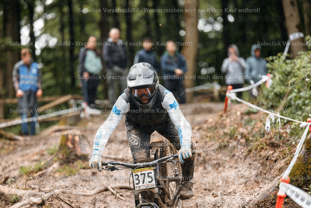 Adrenalin pur: Mountainbiker im Full Speed | Ein Mountainbiker rast einen anspruchsvollen Trail hinunter, umgeben von dichtem Wald. Der Fahrer ist in voller Schutzausrüstung, einschließlich Helm und Goggles, und konzentriert sich voll und ganz auf die Fahrt. Der Trail ist staubig und holprig, mit Wurzeln und Steinen, die die Fahrt erschweren. Im Hintergrund sind Zuschauer zu sehen, die den Fahrer anfeuern. - Realisiert mit Pictrs.com