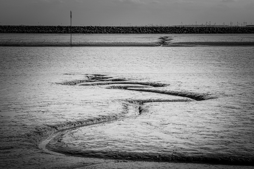 Wasserrinne | Eine Wasserrinne bei Ebbe im Schlick der Nordsee, der auf einen Priel zuläuft, hinter dem sich eine Lahnung aus großen Steinen befindet. — Auflösung des Originals: 6015 x 4010 px. - Realisiert mit Pictrs.com