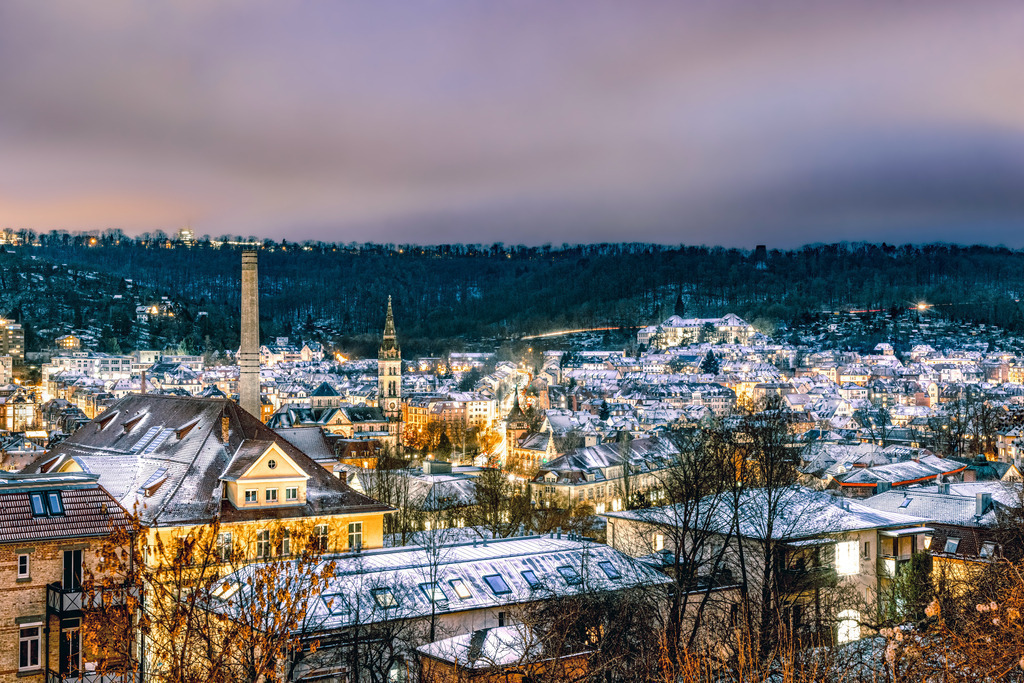 Blick über das winterliche Stuttgart Heslach bei Nacht,Schnee | Dies ist eine winterliche Nachtaufnahme der Stadt Stuttgart in Deutschland. Das Bild zeigt die Landeshauptstadt Baden-Württembergs bei Nacht, bedeckt mit Schnee.Die Stadtbeleuchtung sorgt für einen warmen Kontrast zur kalten, schneebedeckten Landschaft.Im Hintergrund sind bewaldete Hügel zu sehen, die ebenfalls leicht angezuckert sind.Auffällig sind ein hoher Schornstein und eine Kirchturmspitze, die aus der Skyline herausragen. - Realisiert mit Pictrs.com