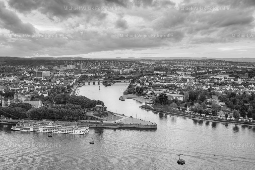 Blick von der Festung Ehrenbreitstein auf das Deutsche Eck in Koblenz | Das Foto zeigt den Blick von der Festung Ehrenbreitstein auf das Deutsche Eck in Koblenz. In Schwarz-Weiß gehalten, wirkt die Szene reduziert und klar. Die Wolken über dem Zusammenfluss von Rhein und Mosel sind dicht und kontrastreich, was dem Bild eine gewisse Spannung verleiht. Das Reiterstandbild von Wilhelm I. ist gut zu erkennen, eingebettet in die markante Flusslandschaft. Eine ruhige, sachliche Aufnahme mit starker Perspektive. - Realisiert mit Pictrs.com