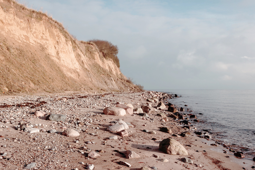 Wandbild: Morgenstimmung an der Steilküste | Dieses Wandbild zeigt die Steilküste am Meer in dezenten Farben. Die Abbruchkante der Steilküste ist beige und braun. Zwischen der Steilküste und dem Meer befindet sich ein Naturstrand an dem viele Steine liegen. Schaffen Sie sich ein maritimes Ambiente in Ihrem Wohnzimmer und holen Sie sich dieses stilvolle Wandbild. Es ist auf Leinwand, Aluminium-Platte, Acrylglas oder als Holzdruck erhältlich. Die Wandbilder werden individuell für Sie in vielen Abmessungen produziert. Daher passen die Ostseekult Wandbilder immer perfekt an Ihre Wände. - Realisiert mit Pictrs.com