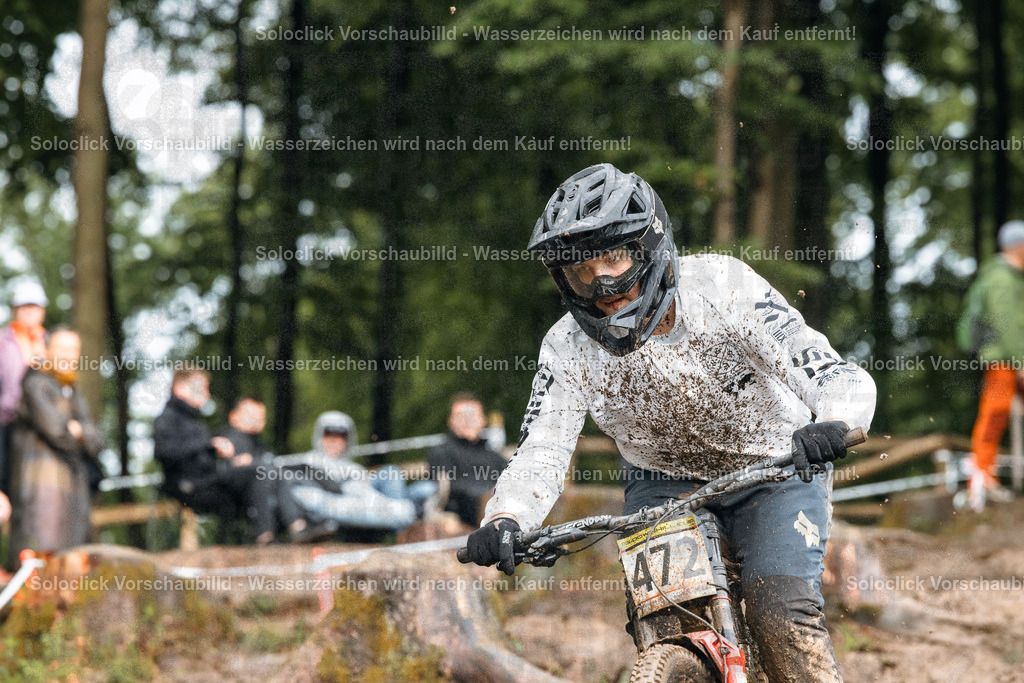 Mountainbike-Action im Wald | Ein Mountainbiker rast mit hoher Geschwindigkeit durch einen staubigen Waldweg. Er trägt einen schwarzen Helm und eine Maske, die sein Gesicht bedecken, sowie ein weißes T-Shirt und schwarze Hosen. Auf seinem Rücken befindet sich eine Nummer 474. Der Hintergrund zeigt einen dichten Wald mit vielen Bäumen und einigen Zuschauern, die am Wegesrand stehen. Der Weg ist mit Erde und Steinen bedeckt, was die Fahrt anspruchsvoll macht. - Realisiert mit Pictrs.com