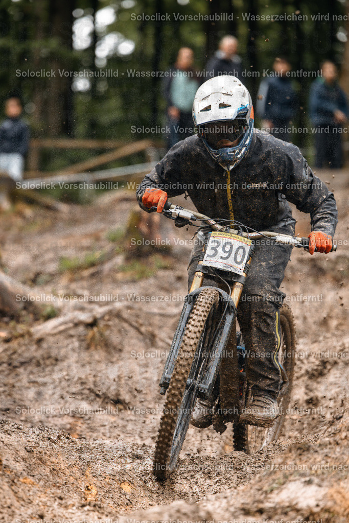 Mountainbike-Action im Schlamm | Das Foto zeigt einen Mountainbiker in voller Ausrüstung, der durch einen schlammigen, unebenen Waldpfad fährt. Der Biker trägt einen weißen Helm mit schwarzen Akzenten und eine dunkle Schutzkleidung. Er fährt ein Full-Suspension-Mountainbike mit breiten Reifen, das offensichtlich für anspruchsvolles Gelände geeignet ist. Der Pfad ist mit Schlamm bedeckt und weist Wurzeln und Steine auf. Im Hintergrund sind Bäume und andere Zuschauer zu sehen. - Realisiert mit Pictrs.com