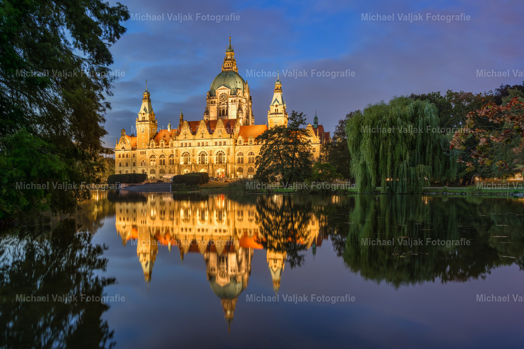 Abendstimmung am Neuen Rathaus in Hannover | Im sanften Licht der Dämmerung erhebt sich das Neue Rathaus majestätisch am Ufer des Maschteichs. Die kunstvoll beleuchtete Fassade spiegelt sich klar im ruhigen Wasser und verleiht der Szene eine fast märchenhafte Atmosphäre. Rechts im Bild rahmt eine Trauerweide mit ihren herabhängenden Zweigen das Ensemble und unterstreicht die stille Eleganz des Moments. Ein Bild, das Architektur und Natur in harmonischer Balance zeigt – eindrucksvoll und zugleich beruhigend. - Realisiert mit Pictrs.com