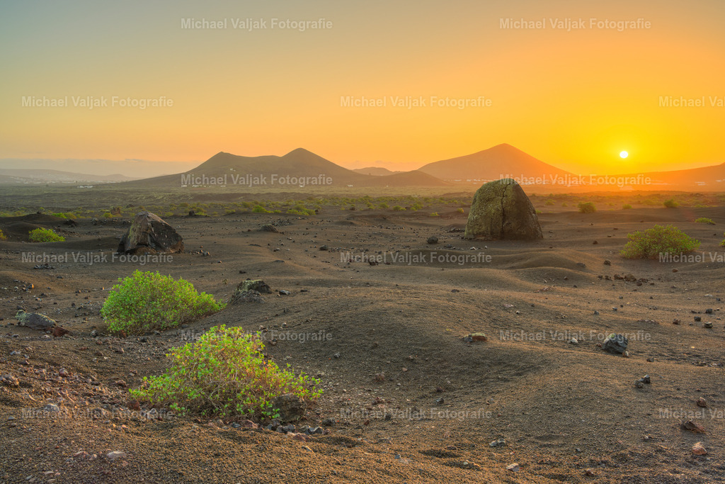 Vulkanlandschaft auf Lanzarote im Zauber des ersten Sonnenlichts | Die mächtige Vulkanbombe am Montana Colorada prägt das Bild, während im Hintergrund der Volcán de Juan Bello, die Häuser von Masdache und der Montana Blanca sichtbar werden. Die Sonne ist gerade aufgegangen und erscheint als helle Kugel im morgendlichen Dunst - ein Moment, in dem die karge Vulkanlandschaft und das warme Licht eine eindrucksvolle Szenerie formen. - Realisiert mit Pictrs.com