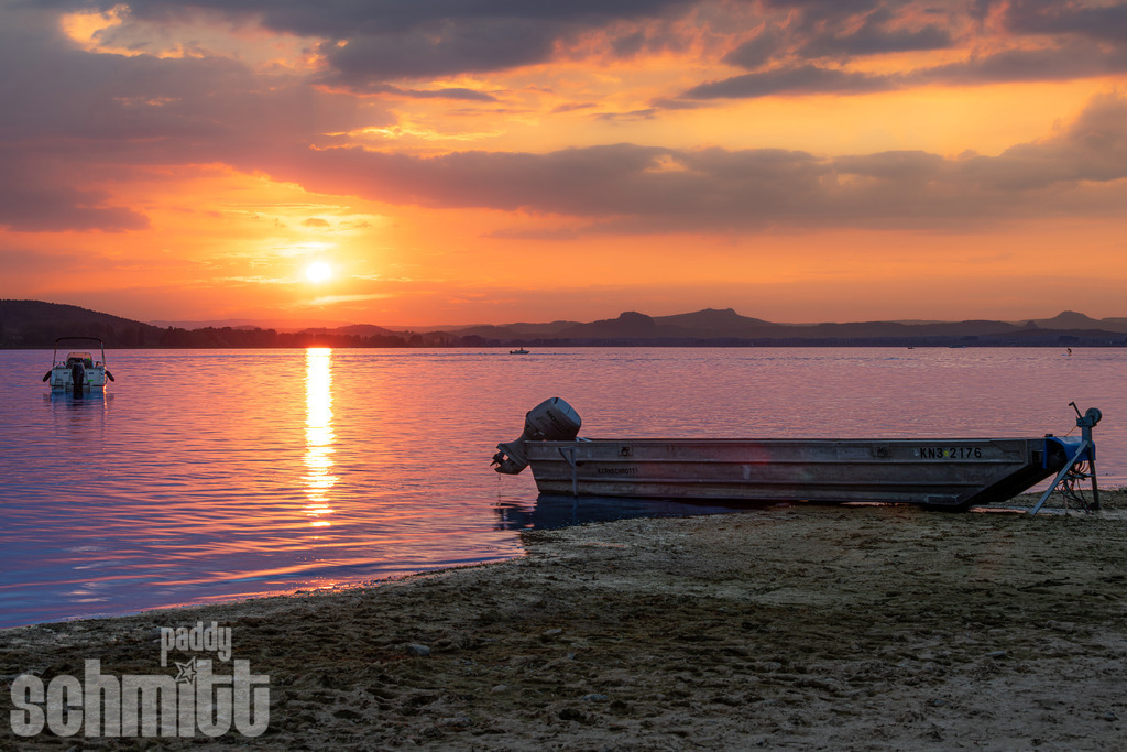 Sonnenuntergang am Seeufer bei der Insel Reichenau am Bodensee | Am Seeufer bei der Insel Reichenau taucht die untergehende Sonne den Bodensee in warme, goldene Farben. Das ruhige Wasser, der leuchtende Horizont und die sanfte Abendstimmung schaffen eine besonders idyllische und stimmungsvolle Szenerie. - Realisiert mit Pictrs.com