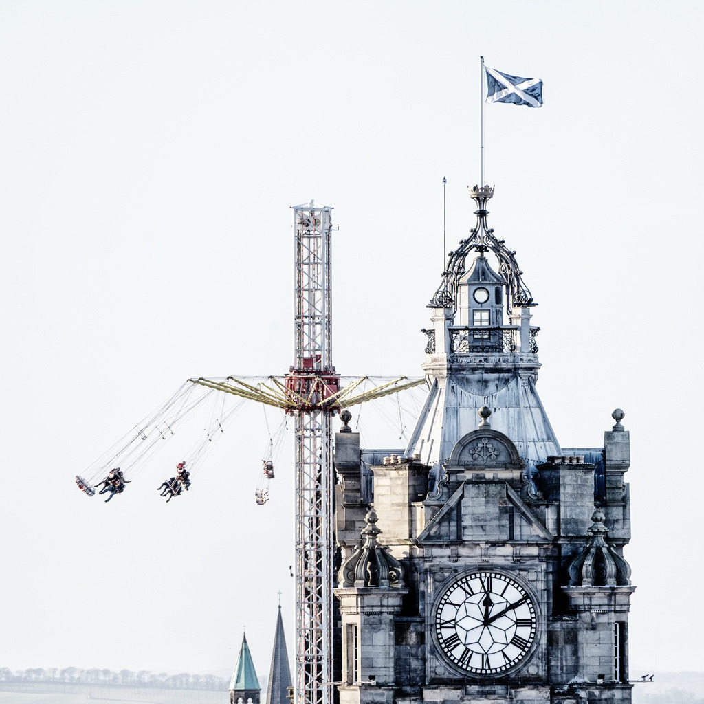Edinburgh: Kontrast am Balmoral Hotel | Ein historischer Uhrenturm aus hellem Sandstein, gekrönt von einer schmiedeeisernen Kuppel und der blau-weissen schottischen Flagge (Saltire), steht im Kontrast zu einem modernen Kettenkarussell direkt daneben. Die Gondeln des Fahrgeschäfts hängen hoch in der Luft, während Personen in schwindelerregender Höhe über der Stadt kreisen. Die Zifferblätter der grossen Turmuhr zeigen kurz nach zwölf. Der Hintergrund ist in ein helles, fast weisses Licht getaucht, was die Silhouette der Architektur und das filigrane Stahlgerüst des Karussells scharf hervorhebt.***### 10 tags[Edinburgh, Balmoral Hotel, Clock Tower, Winter Festival, Christmas Market, Swing Ride, Scottish Flag, Contrast, Old vs New, Dynamic] - Realisiert mit Pictrs.com