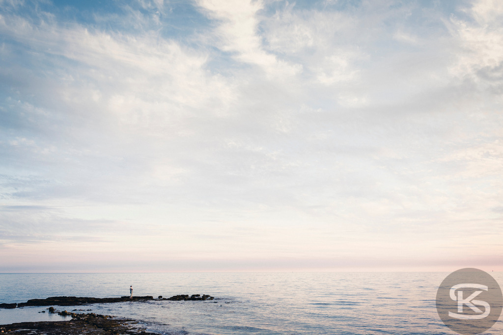 Ruhige Meereslandschaft bei Dämmerung, einsame Person auf Felsen | Ein friedliches und minimalistisches Bild einer ruhigen Meereslandschaft während der Dämmerung. Die pastellfarbenen Wolken in Hellblau und Rosa treffen auf den Horizont, während eine kleine, einsame Silhouette auf einem Felsen im Vordergrund steht. Das Bild strahlt Ruhe, Gelassenheit und Harmonie aus. - Realisiert mit Pictrs.com