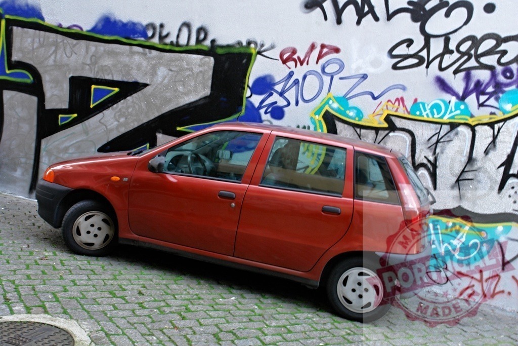 Red car parked on urban cobblestone street with graffiti wall | Red compact car parked on a steep cobblestone street featuring colorful street art on the wall - Realisiert mit Pictrs.com