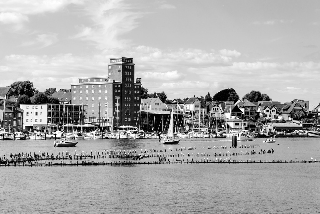 Wandbild: Hafen in Kappeln in Schwarz-Weiß | Genießen Sie den beeindruckenden Anblick des Hafens in Kappeln mit dieser faszinierenden Schwarz-Weiß-Aufnahme. Das Bild zeigt moderne und traditionelle Gebäude, die den Charme und die Geschichte der Stadt widerspiegeln. Auf der Schlei befinden sich elegante Segelboote, die dem Bild eine maritime Note verleihen.Dieses Kunstwerk, erhältlich auf Leinwand, Alu-Dibond, Acrylglas, Fineart und als Akustikbild, bringt die lebendige Atmosphäre und die Schönheit des Hafens von Kappeln direkt in Ihr Zuhause. - Realisiert mit Pictrs.com