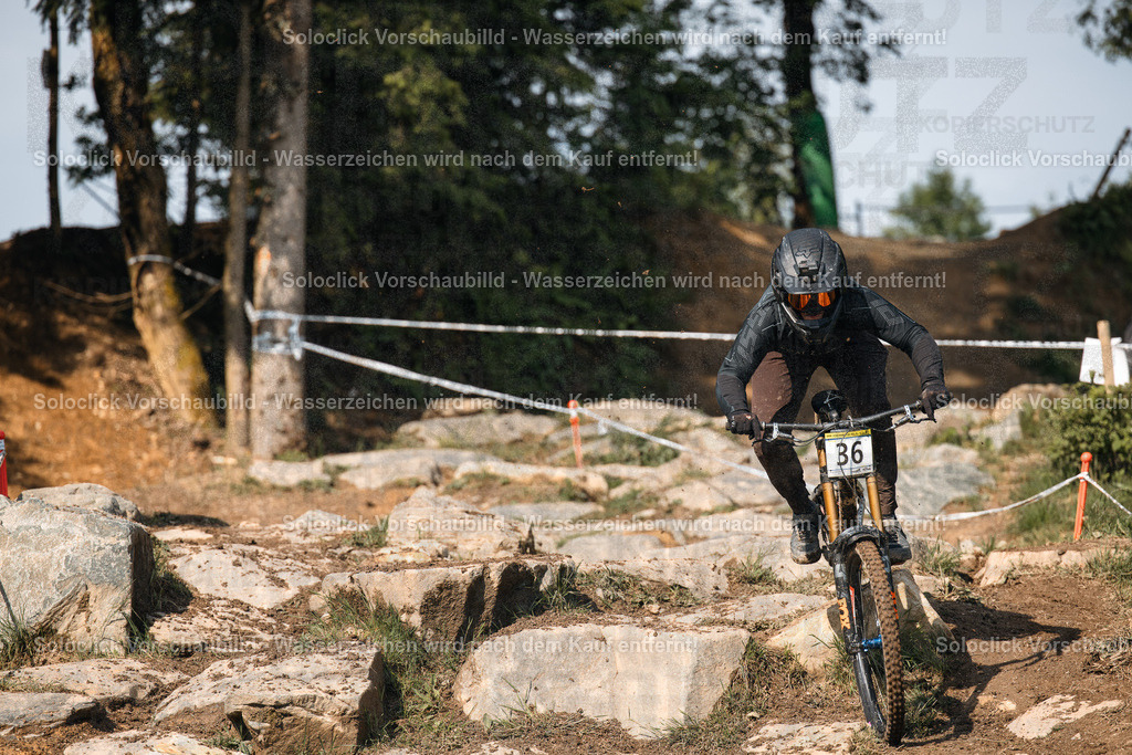 Adrenalin pur: Mountainbiker im Wald | Ein Mountainbiker in voller Ausrüstung rast mit hoher Geschwindigkeit einen steinigen, kurvigen Pfad durch einen dichten Wald hinunter. Er trägt einen schwarzen Helm mit Visier und eine schwarze Jacke mit dem Nummernschild '36'. Der Pfad ist mit großen Steinen und Wurzeln übersät, was die Fahrt anspruchsvoll macht. Im Hintergrund sind weitere Bäume und Büsche zu sehen, die die natürliche Umgebung unterstreichen. - Realisiert mit Pictrs.com