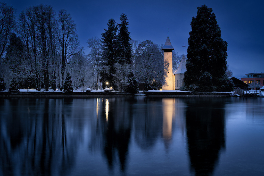 Kirche Scherzligen im Winter | Die beleuchtete Kirche Scherzligen steht majestätisch am Ufer des Thunersees in Thun, umgeben von schneebedeckten Bäumen in der frühen Morgenstunde. Das ruhige Wasser des Sees reflektiert die warmen Lichter der Kirche und die dunklen Silhouetten der Bäume, wodurch eine stimmungsvolle Symmetrie entsteht. Die Szene fängt die friedliche und kalte Schönheit eines Wintermorgens in den Schweizer Alpen ein. - Realisiert mit Pictrs.com
