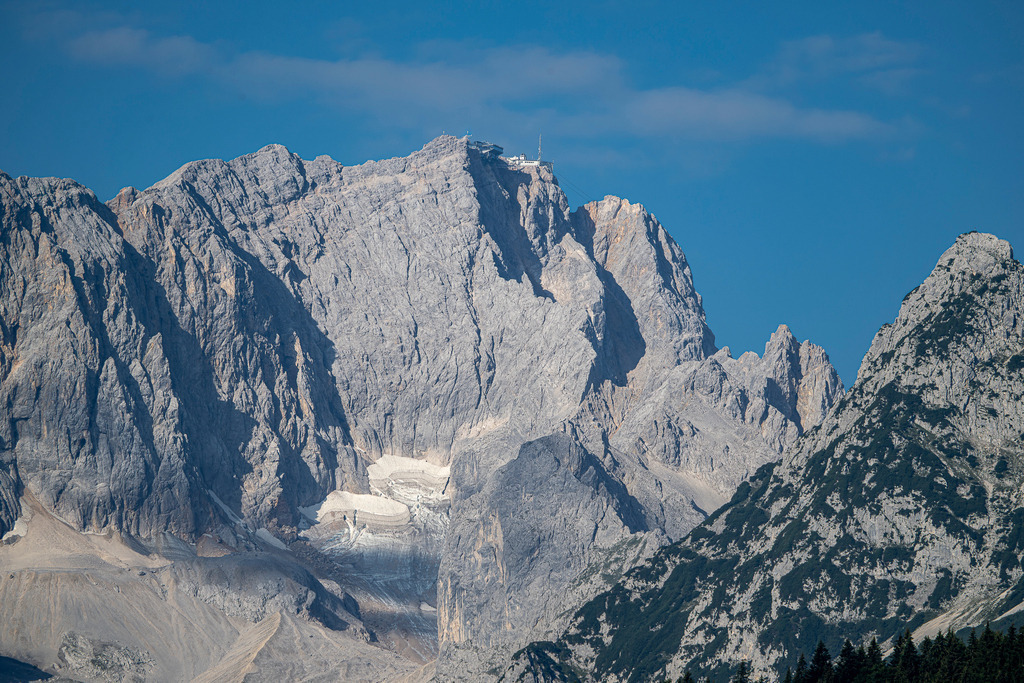 Majestätischer Gipfelblick: Die Zugspitze in ihrer vollen Pracht | Ein imposantes Panorama über Garmisch-Partenkirchen: Glasklar und kraftvoll zeichnet sich das Zugspitzmassiv vor einem strahlend blauen Sommerhimmel ab. Am Gipfel thront die charakteristische Bergstation der Zugspitzbahn, während weiter unten im schattigen Felskessel die schwindenden Reste des Höllentalferners zu erkennen sind. Die markanten Kalkfelsen und schroffen Gradverläufe spiegeln die wilde Schönheit des Wettersteingebirges wider – eine eindrucksvolle Aufnahme voller Erhabenheit, alpiner Faszination und Naturkraft. - Realisiert mit Pictrs.com
