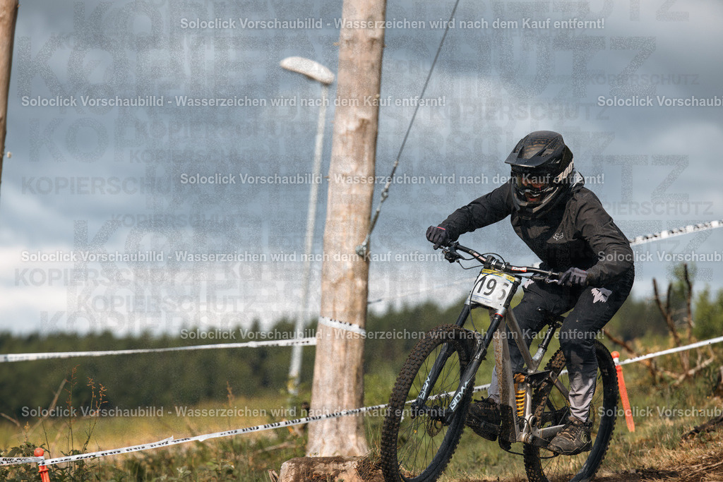 Action im Bikepark: Downhill-Fahrer im iXS Downhill Cup | Das Foto zeigt einen Mountainbiker während des iXS Downhill Cups 2025 im Bikepark Winterberg, speziell auf der Strecke 'The Mother'. Der Fahrer ist in voller Montur unterwegs und bewältigt mit hoher Geschwindigkeit eine kurvige Passage. Der Hintergrund besteht aus Bäumen und einer staubigen Strecke, die mit Absperrband markiert ist. Der Himmel ist bewölkt und deutet auf möglicherweise feuchtes Wetter hin. - Realisiert mit Pictrs.com