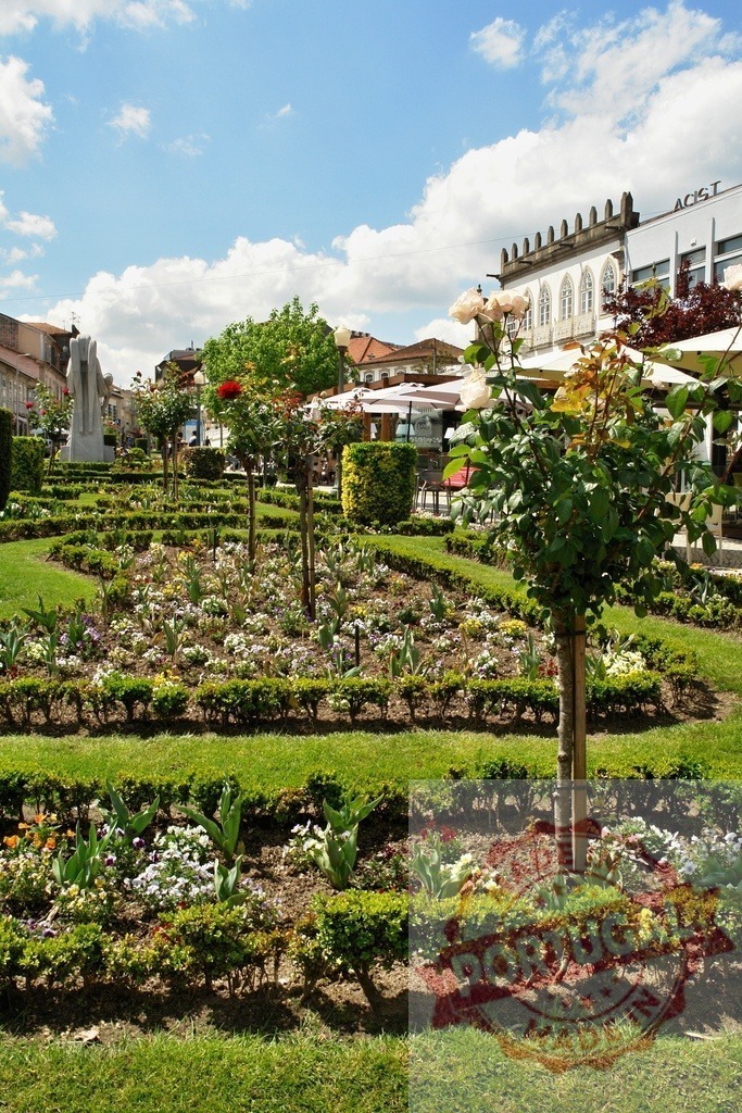 Public garden with flowers and building under blue sky. Region Norte - Portugal | Manicured garden beds displaying flowering plants and green hedges with a town square in the background - Realisiert mit Pictrs.com