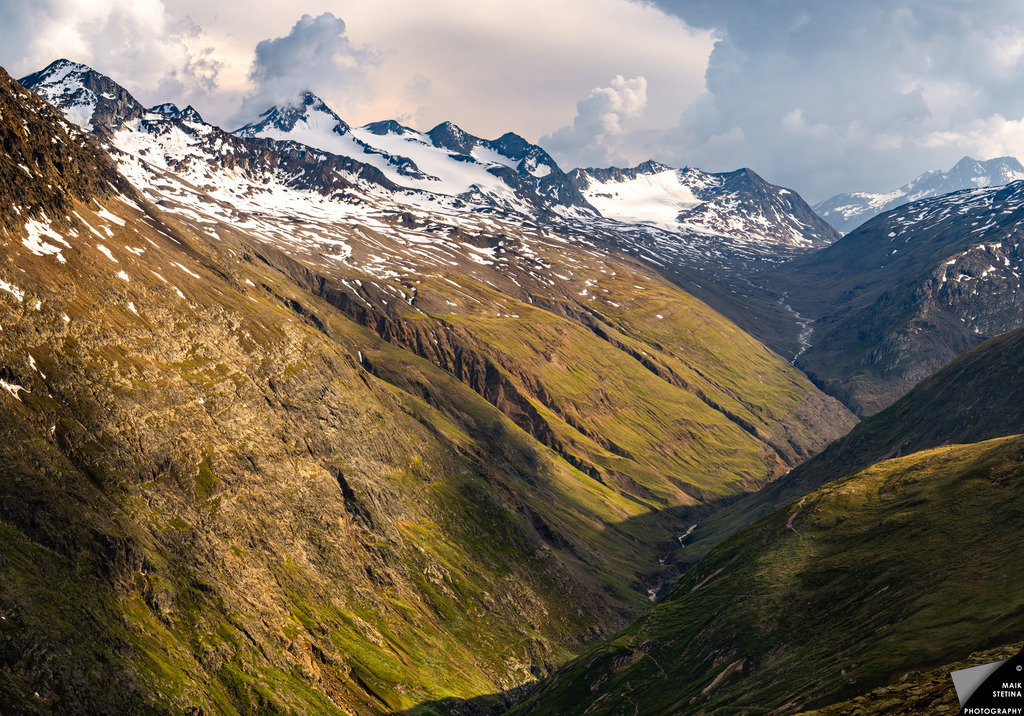 Stormlight Over the High Alpine Valleys from Breslauer Hut | From the Breslauer Hut at around 2,900 meters, I looked out across the high alpine valleys and felt the scale of the journey ahead of me. The first thunderstorms had already started building from the direction of Italy, while the setting sun in the west broke through gaps in the clouds and painted the mountains with warm, dramatic light. Below me, the valleys seemed to reveal the route I would follow over the coming days, leading deeper into the Austrian Alps. On the horizon, I could see the Gravwand — a distant ridge that I would cross a few days later — and in that moment the hike felt both vast and deeply personal. - Realisiert mit Pictrs.com