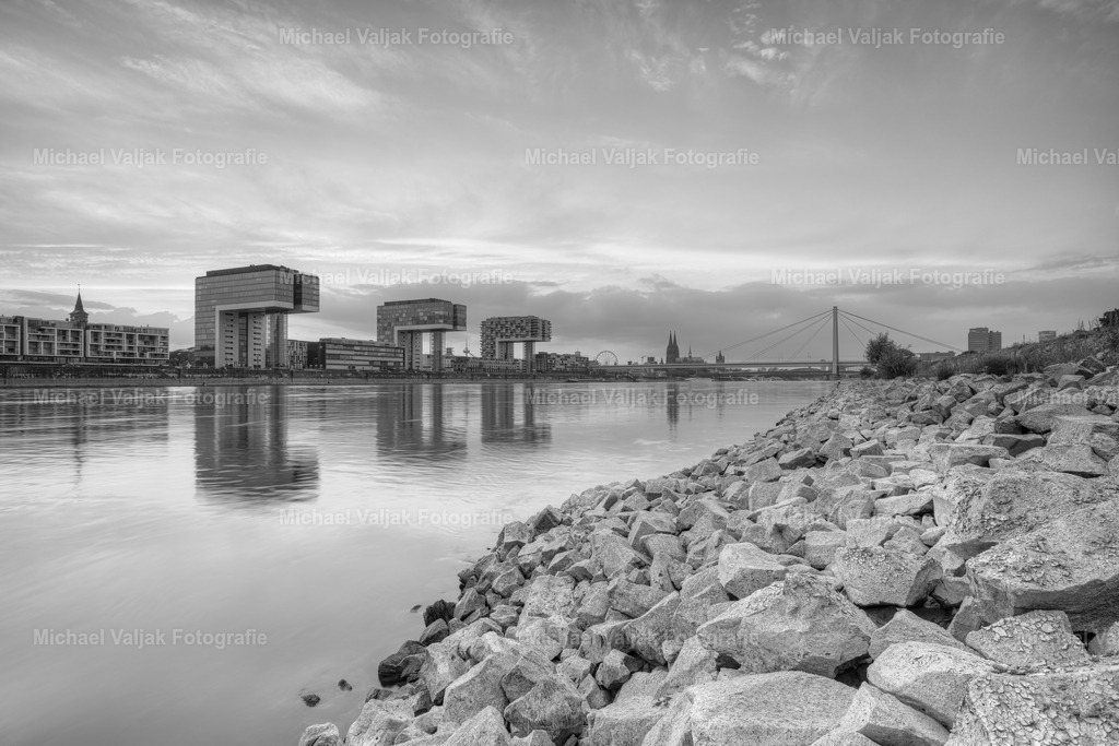Am Rheinufer in Köln schwarz-weiss | Blick von den Pollerwiesen in Richtung Kranhäuser und Kölner Dom im Hintergrund. Der Rhein führt sehr wenig Wasser, bei Normalpegel sind die Steine komplett vom Wasser bedeckt. Die Gebäude der Kölner Skyline spiegeln sich im relativ ruhigen Wasser. - Realisiert mit Pictrs.com