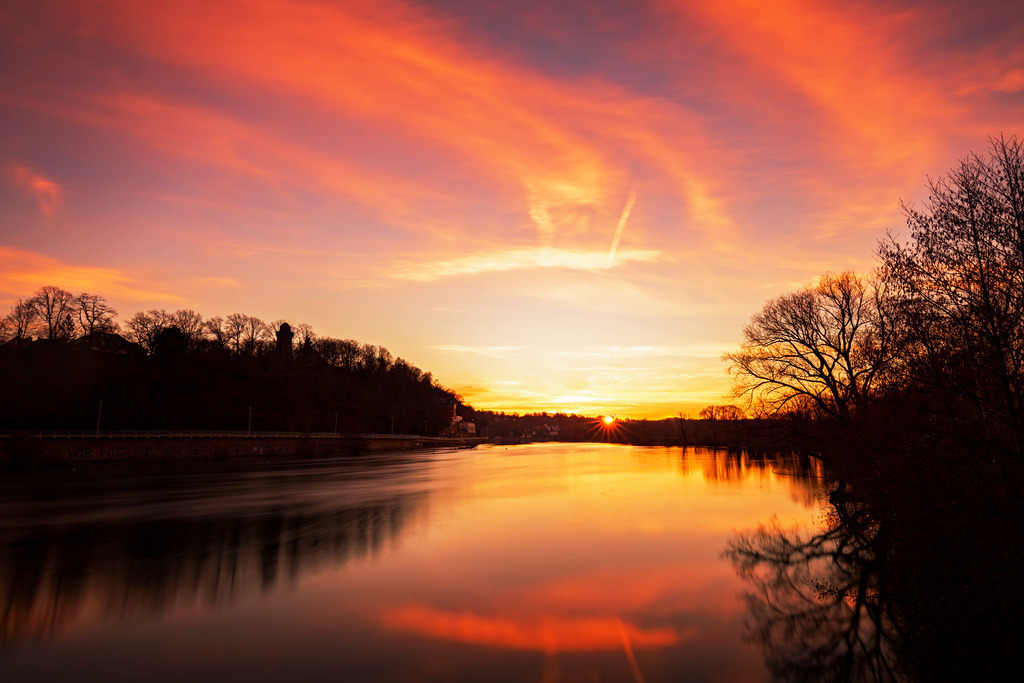 Wandbild: Magischer Sonnenuntergang am Fluss | Dieses Wandbild fängt die ruhige Schönheit eines Sonnenuntergangs an einem Flussufer ein. Der Himmel erstrahlt in leuchtenden Orange- und Rosatönen, die sich harmonisch im stillen Wasser des Flusses widerspiegeln. Die Silhouetten der Bäume und die sanften Hügel im Hintergrund verleihen der Szenerie eine friedliche und doch dramatische Atmosphäre. Das Bild vermittelt ein Gefühl der Ruhe und Gelassenheit und bringt die Magie eines Sonnenuntergangs direkt in Ihr Zuhause.