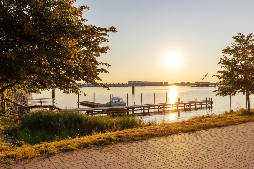 Wandbild: Sonnenaufgang an der Promenade in Olpentiz | Dieses Wandbild im Querformat zeigt einen schönen Sonnenaufgang an der Promenade in Olpenitz. Auf dem Wasser befindet sich ein Bootsanleger. Die morgendliche Sonne steht tief am Horizont. Auf dem Wasser ergibt sich eine schöne Sonnenreflexion. Der leicht rötliche Himmel ist wolkenlos. - Realisiert mit Pictrs.com