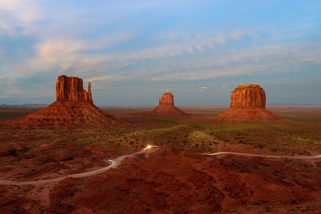usa-2015-294 | Panorama mit den Tafelbergen West Mitten Butte, East Mitten Butte und Merrick Butte (von links nach rechts) im Monument Valley (USA) nach Sonnenuntergang. Die letzten Fahrzeuge verlassen Monument Valley und zeichnen mit ihren Scheinwerfern aufgrund der langen Belichtungszeit ein Muster in die Landschaft. - Realisiert mit Pictrs.com