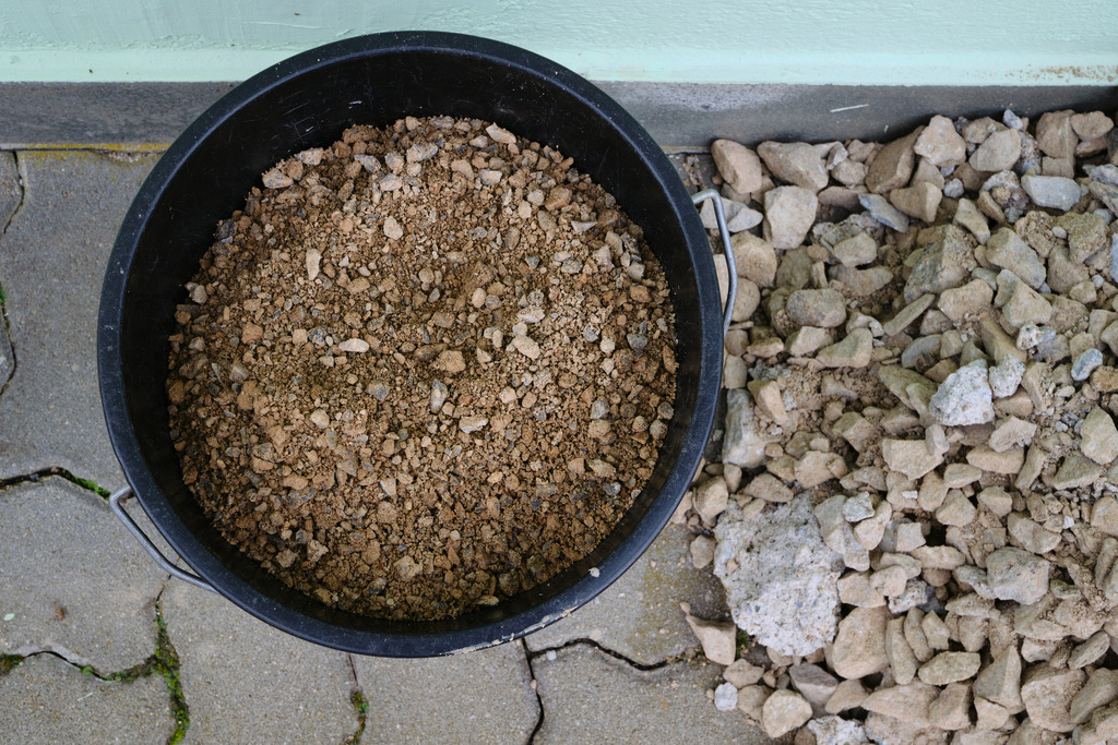 Schwarzer Eimer mit Kiesfüllung auf dem Weg | Austria - October 08, 2023: Schwarzer Eimer mit Kiesfüllung auf dem Weg, daneben ein Steinhaufen. - Realisiert mit Pictrs.com