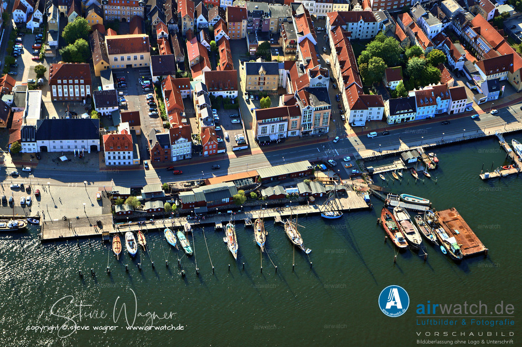 Luftbild Flensburger Foerde, Flensburg Binnenhafen, Schiffbrücke, Museumswerft | Die Museumswerft ist eine gemeinnützige GmbH, die von einem Förderverein durch ehrenamtliche Arbeit, Mitgliedsbeiträge und Spenden unterstützt wird. Sie zeigt und baut traditionelle Segel- und Arbeitsboote, wie sie vor 100 bis 200 Jahren im Ostseeraum im Handel und in der Fischerei unterwegs waren. Besonders hervorgehoben wird die Restaurierung und Nachbildung historischer Schiffe, wie beispielsweise einer Drei-Masten-Brigantine aus dem Jahr 1781, die ursprünglich in der Nähe des heutigen Standorts vom Stapel lief.