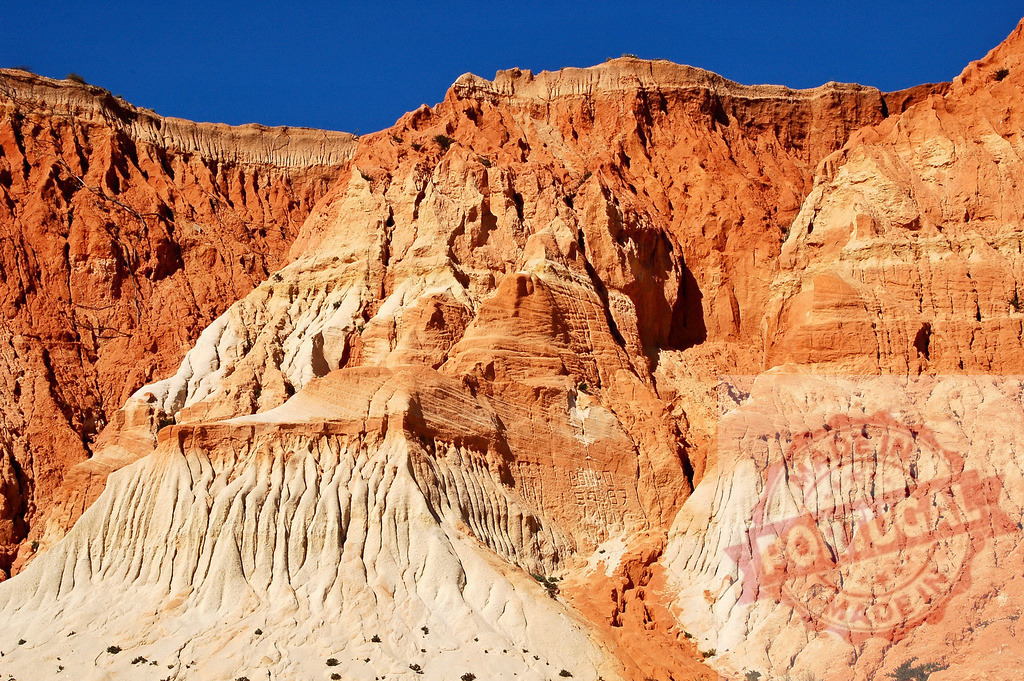 Layered red and white sandstone cliffs experiencing erosion in Algarve | Red and white sandstone cliffs showing natural erosion patterns against a bright blue sky - Realized with Pictrs.com