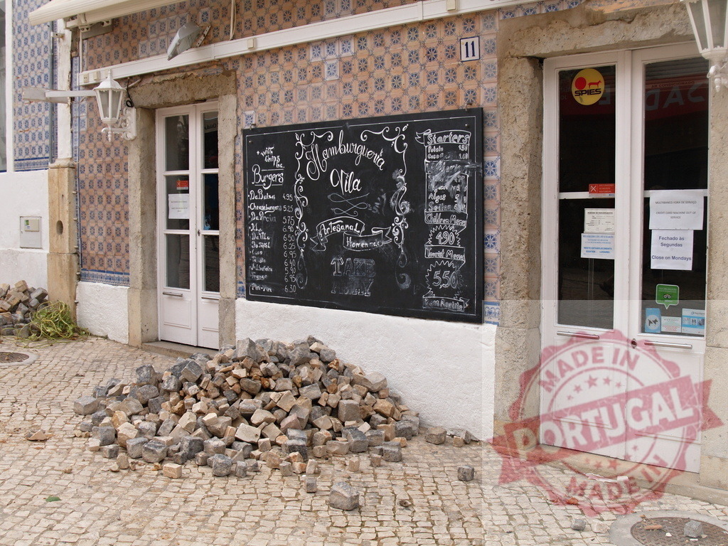 Portuguese restaurant facade undergoing street repair work | Albufeira Flood - discruction by weather problems Cobblestones piled in front of a Portuguese restaurant with azulejo tiles and a chalkboard menu - Realisiert mit Pictrs.com