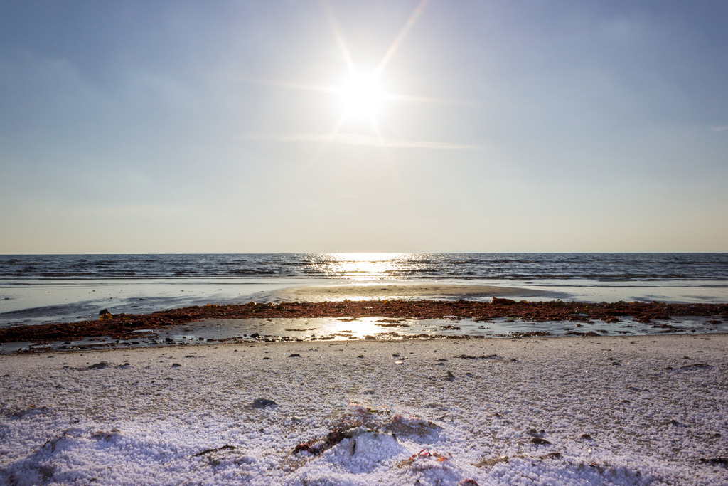 Wandbild: Frostiger Morgen am sonnigen Sandstrand | Dieses Wandbild im Querformat zeigt eine schöne Morgenstimmung im Winter am Meer. Im Vordergrund ist der leicht verschneite Strand zu sehen. Die morgendliche Sonne scheint am blauen Himmel und spiegelt im Meer.  - Realisiert mit Pictrs.com