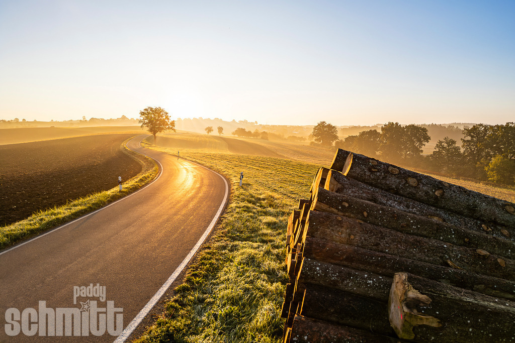 Idyllischer Sonnenaufgang auf dem Land | Ein idyllischer Sonnenaufgang taucht die ländliche Landschaft in warmes, sanftes Licht. Eine ruhige Landstraße führt durch die Natur, während ein Holzstapel am Wegesrand die schöne Atmosphäre der ländlichen Umgebung unterstreicht. - Realisiert mit Pictrs.com