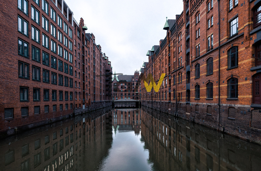 Blick in eine Wasserstraße der Speicherstadt in Hamburg,Backsteingebäude | Das Bild zeigt die Hamburger Speicherstadt. Es handelt sich um den größten zusammenhängenden historischen Lagerhauskomplex der Welt, der zum UNESCO-Weltkulturerbe gehört. Die Speicherstadt wurde zwischen 1885 und 1927 erbaut. Sie liegt südlich der Hamburger Altstadt, zwischen dem Oberhafen und dem Sandtorkai. Die charakteristische Architektur aus roten Backsteingebäuden und Fleeten (Kanälen) spiegelt sich im Wasser wider.  - Realisiert mit Pictrs.com