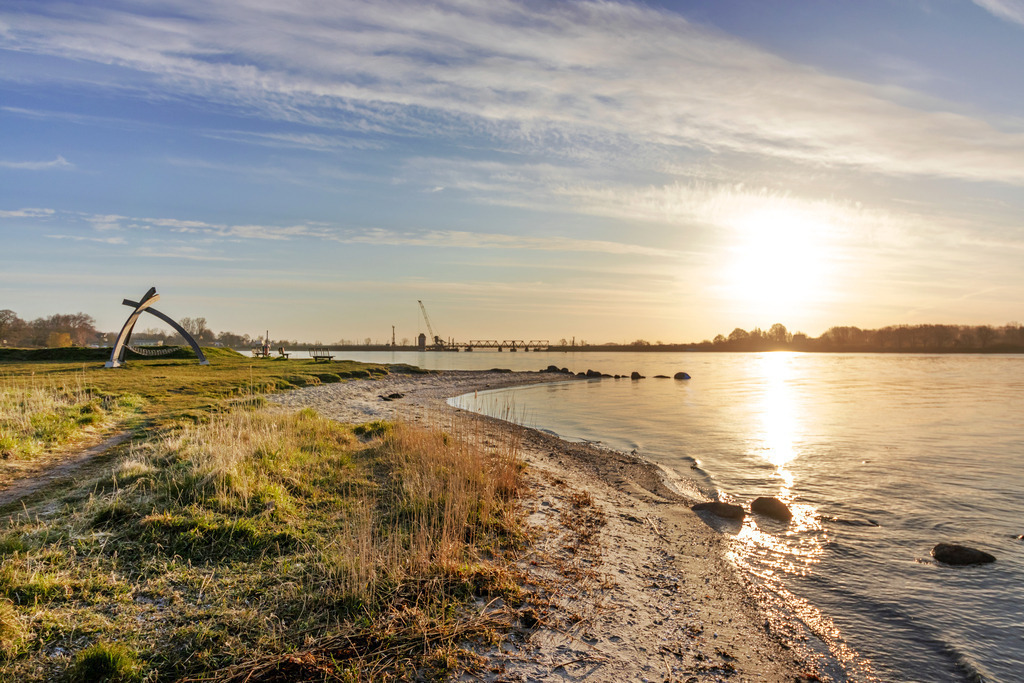 Wandbild: Sonnenaufgang am Schleistrand Schneiderhaken | Dieses Wandbild zeigt den Schleistrand Schneiderhaken in einer schönen morgendlichen Lichtstimmung. Die Sonne befindet sich direkt über der Schlei. Auf der Schlei ergibt sich eine schöne Lichtreflexion. Auf der linken Seite befindet sich der Schleistrand. Direkt am Schleistrand steht ein Netzlausleger. In der Ferne kann man die Schleibrücke in Lindaunis erahnen. - Realisiert mit Pictrs.com