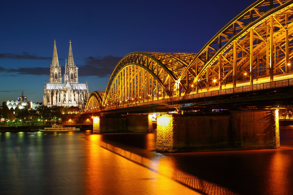 Köln bei Nacht - Glanz der Hohenzollernbrücke | Dieses eindrucksvolle Wandbild fängt die nächtliche Magie der Rheinmetropole ein: Die Hohenzollernbrücke in leuchtendem Gold spannt sich über den ruhig dahinfließenden Rhein, während der Kölner Dom majestätisch und detailreich im Hintergrund thront. Die Spiegelungen im Wasser und die klare Architektur schaffen eine harmonische Verbindung von Geschichte, Technik und Atmosphäre – ideal für Stadtliebhaber, Rheinromantiker und Fans urbaner Eleganz.