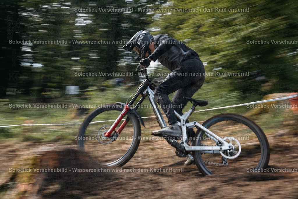 Rasante Fahrt | Ein Mountainbiker in voller Ausrüstung rast mit hoher Geschwindigkeit einen staubigen Pfad hinunter. Die Bewegung ist durch die lange Belichtungszeit deutlich sichtbar, was einen dynamischen Effekt erzeugt. Der Fahrer trägt einen schwarzen Helm und eine schwarze Jacke, sowie schwarze Handschuhe und Hosen. Das Mountainbike ist mit roten Federgabeln und breiten Reifen ausgestattet. - Realisiert mit Pictrs.com