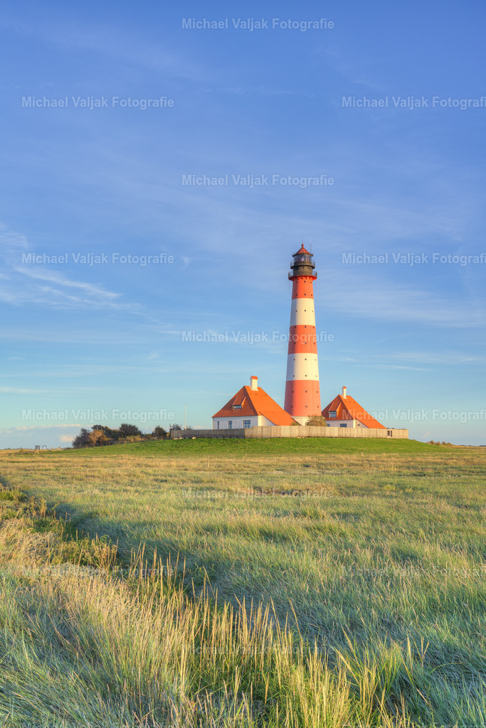 Leuchtturm Westerheversand in der Abendsonne | Der Leuchtturm Westerheversand, ein Wahrzeichen der Halbinsel Eiderstedt in Schleswig-Holstein, steht majestätisch auf einer Warft vor Westerhever. Erbaut im Jahr 1906, hat dieser Leuchtturm eine Feuerhöhe von 41 Metern und dient als See-, Quermarken- und Leitfeuer. Mit seiner imposanten Erscheinung und der Fähigkeit, sein Licht bis zu 21 Seemeilen weit zu tragen, bietet er nicht nur eine lebenswichtige Orientierung für Seefahrer, sondern ist auch ein beliebtes Fotomotiv, besonders in der Abendsonne, welche den Turm in ein goldenes Licht taucht. - Realisiert mit Pictrs.com