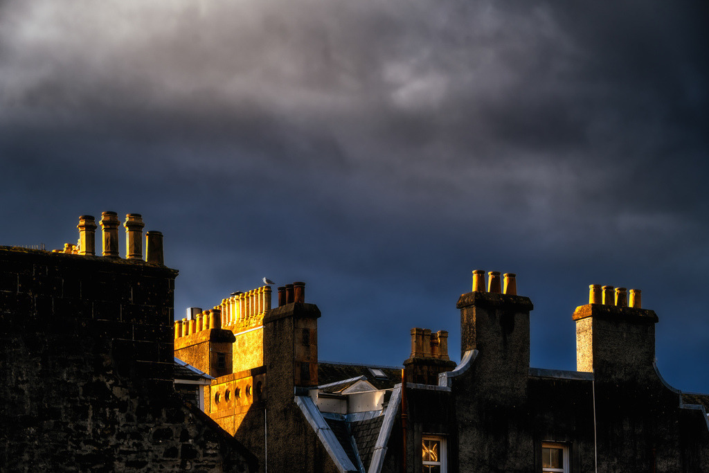 Goldenes Licht über Oban, Schottland | Die Dächer und Schornsteine historischer Gebäude in Oban, Schottland, werden von einem tiefstehenden Sonnenlicht dramatisch beleuchtet. Ein dunkler, bewölkter Himmel bildet einen starken Kontrast zu den goldenen Akzenten auf den Strukturen. Die Komposition betont die architektonischen Details und die atmosphärische Beleuchtung der Stadtlandschaft. - Realisiert mit Pictrs.com
