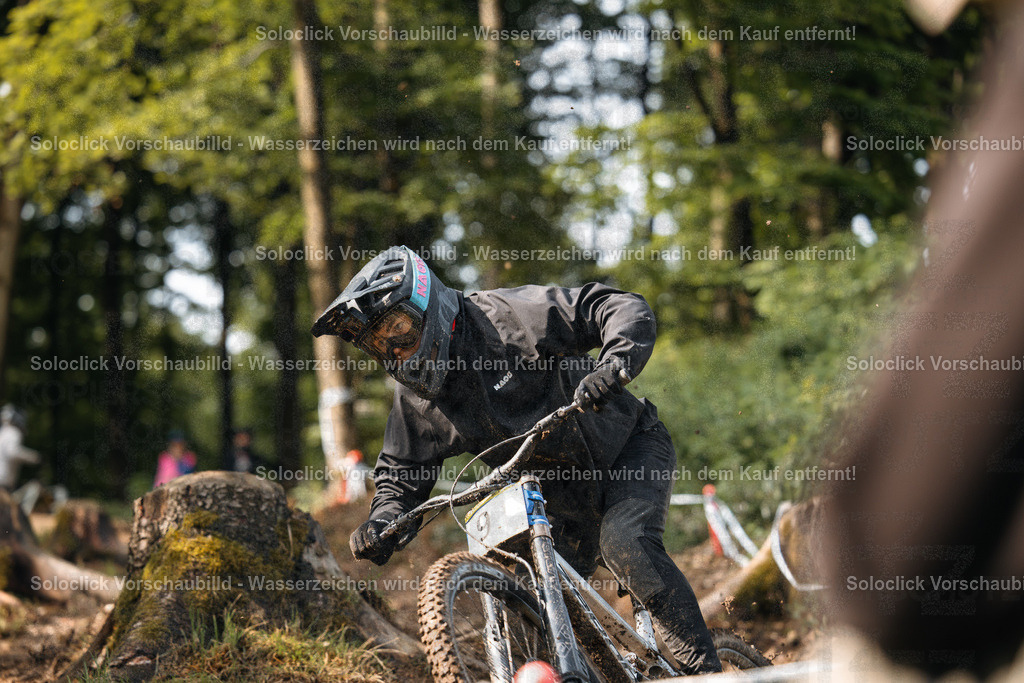 Mountainbike-Action im Wald | Das Foto zeigt einen Mountainbiker in voller Ausrüstung, der einen staubigen, kurvigen Trail in einem bewaldeten Gebiet durchquert. Der Fahrer ist in eine schwarze Jacke und Hose gekleidet und trägt einen schwarzen Helm mit Visier und eine Schutzbrille. Er fährt ein Mountainbike mit Federgabel und breiten Reifen. Der Trail ist staubig und kurvig, und der Hintergrund besteht aus dichtem Grünzeug. Das Bild vermittelt ein Gefühl von Geschwindigkeit und Abenteuer. - Realisiert mit Pictrs.com